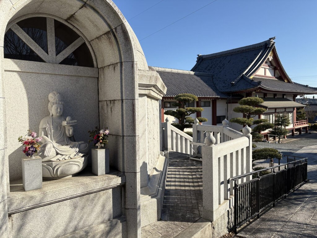 紫雲寺境内の観音菩薩像｜アーチ型の御影石の祠に安置された大悲観音菩薩（蓮の蕾を持つ）・献花・朱色柱の紫雲寺本堂が見える
