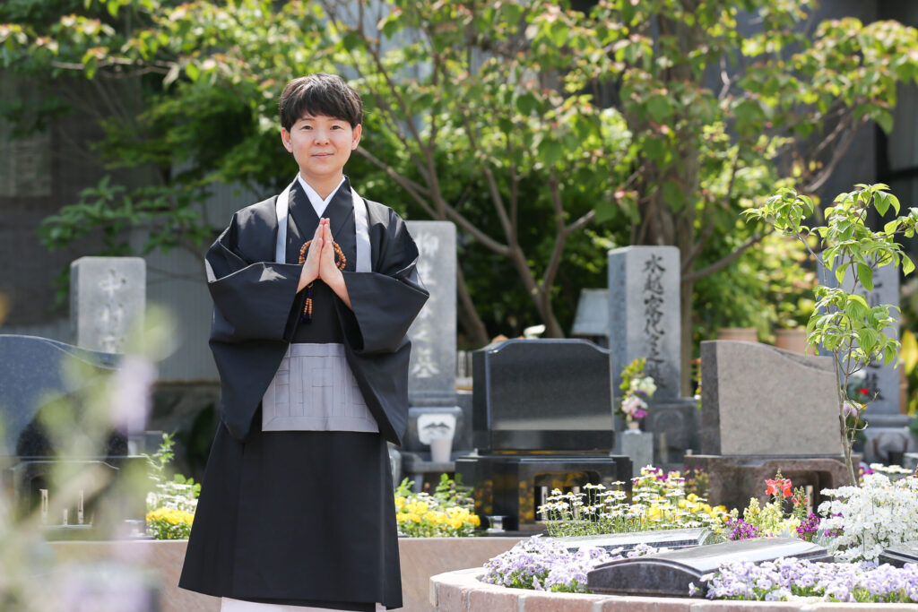 住職 伊藤 春道　専修院 女性住職 尼僧 黒い法衣 グレー袈裟 合掌 念珠 円形樹木葬区画 「中村家之墓」「水越家代々」 紫のビオラ ハナミズキ若木 新緑の境内
