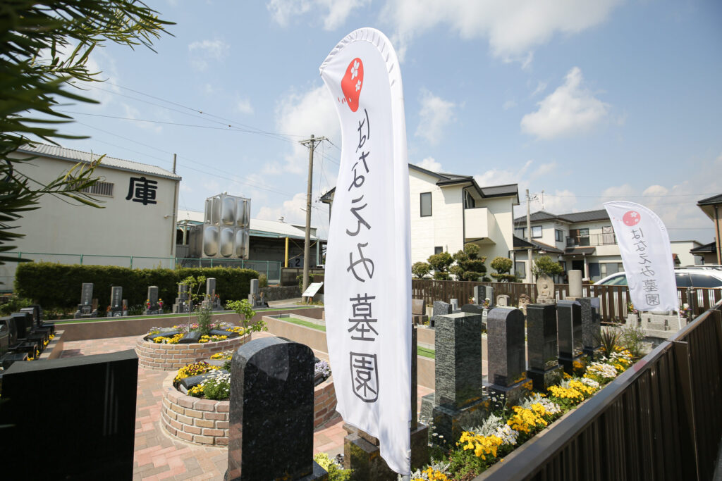 豊橋下地はなえみ墓園 はなえみ墓園フラッグ2本 白地に赤い梅花マーク 円形レンガ樹木葬区画 黄色パンジーの花壇 既存墓地と石仏 青空白雲