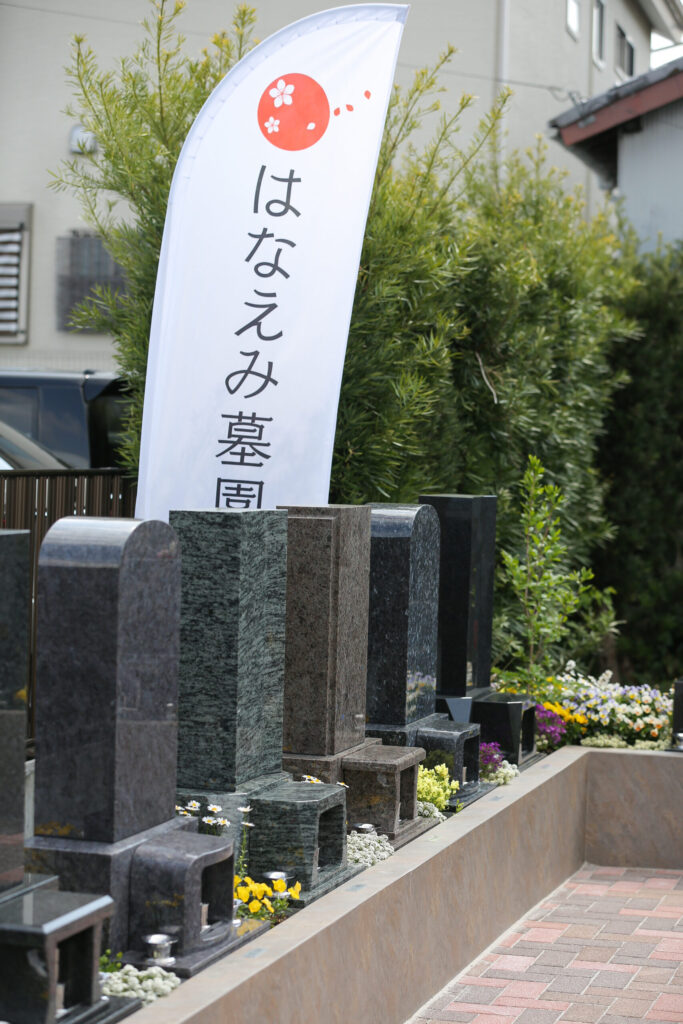 豊橋下地はなえみ墓園 はなえみ墓園フラッグ 白地 赤い梅花マーク 縦書き 縦型洋型墓石ライン 黒・グレー・青影系御影石 ベージュ石製基壇 マキ類常緑生垣 黄色パンジー 白マーガレット