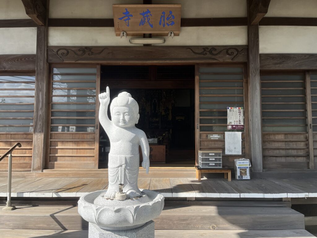 胎蔵寺 本堂正面に建つ石製誕生仏｜右手を天に挙げ「天上天下唯我独尊」の姿、本堂軒下に山号額