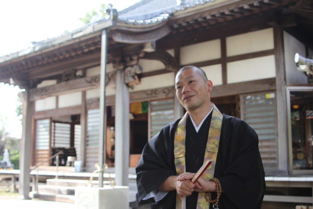 胎蔵寺 住職｜黒い法衣と金襴の輪袈裟・数珠と経本を手に本堂前に立つ住職の姿